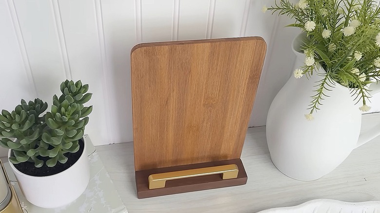 Wooden cookbook holder made from a cutting board and a drawer handle sitting on a table near plants