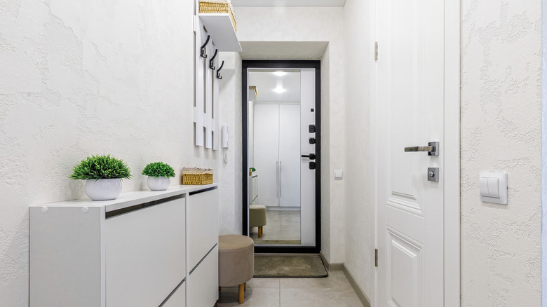 A long, narrow white entryway hallway with a thin table with cabinets, a storage ottoman, and a shelf with hooks hanging on the wall.