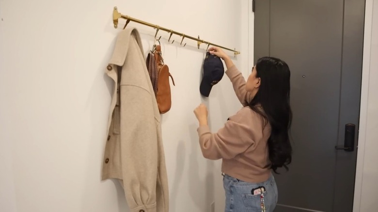 A woman hangs a hat on an entryway rail holding a bag and jacket.