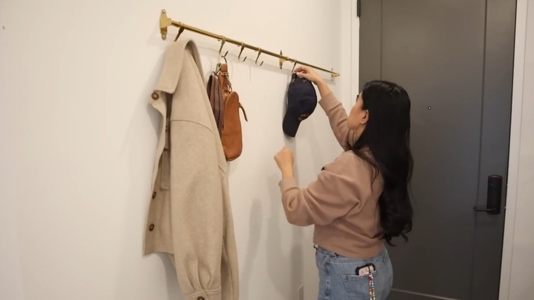 A woman hangs a hat on an entryway rail holding a bag and jacket.