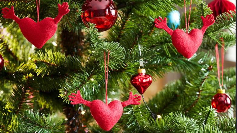 Three cloth hearts with arms hanging from a tree with other red decorations on a Christmas tree