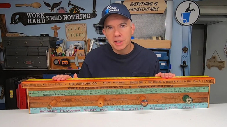 Man shows a coatrack using upcycled yardsticks with wooden spools instead of hooks.