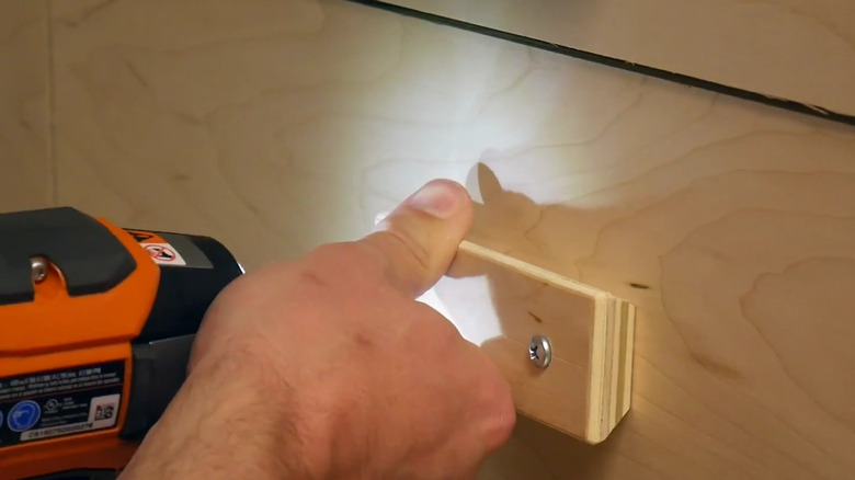 A person uses a drill to screw in a handmade wooden drawer pull that is rectangular in shape with one screw on each end of the horizontal handle.
