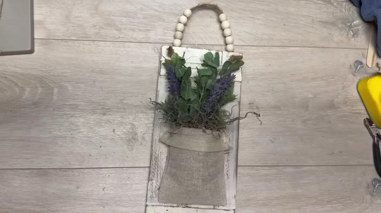 A crafter showcases how she makes a floral pocket hanger using wooden rulers from Dollar Tree as the backdrop, which she stains and distresses before adding a canvas bag for a pocket and dried flowers and other decorations.