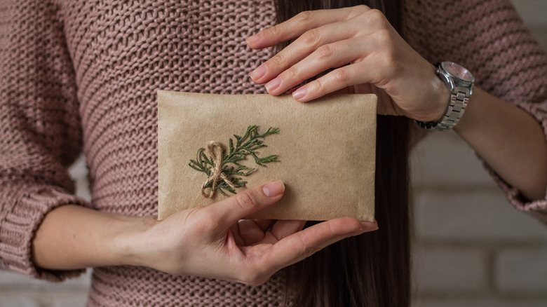 A person holding a brown paper wrapped gift decorated with a sprig of arborvitae