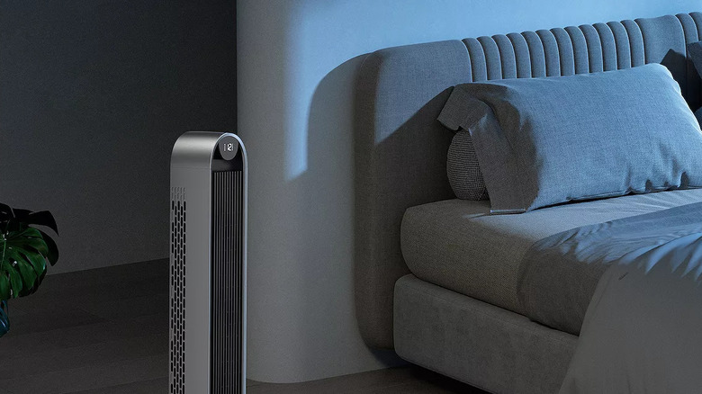 A Dreo Oscillating Tower Fan sits next to a gray bed