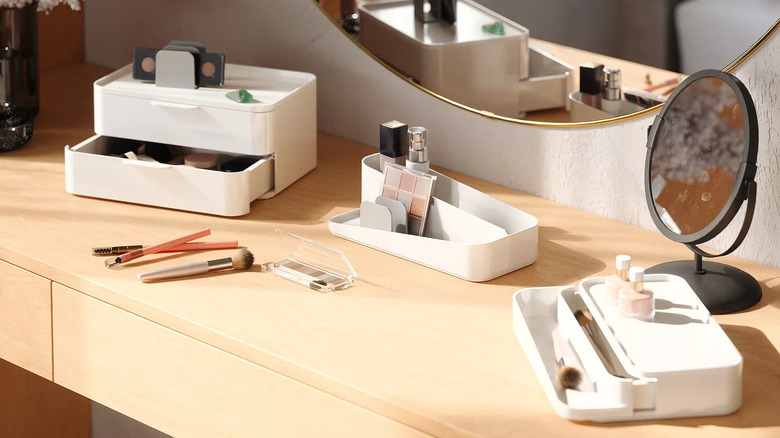 A white tiered cosmetic organizer sits in separate pieces on a wooden vanity beneath a large round mirror