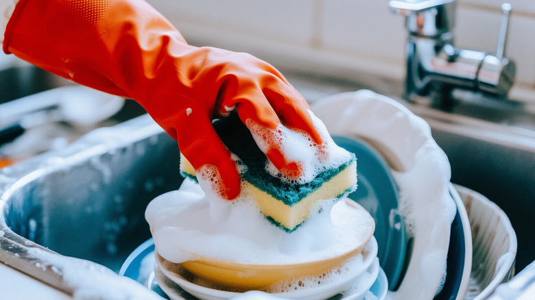 A close-up captures a person washing dishes in a kitchen sink with a sponge and dish soap