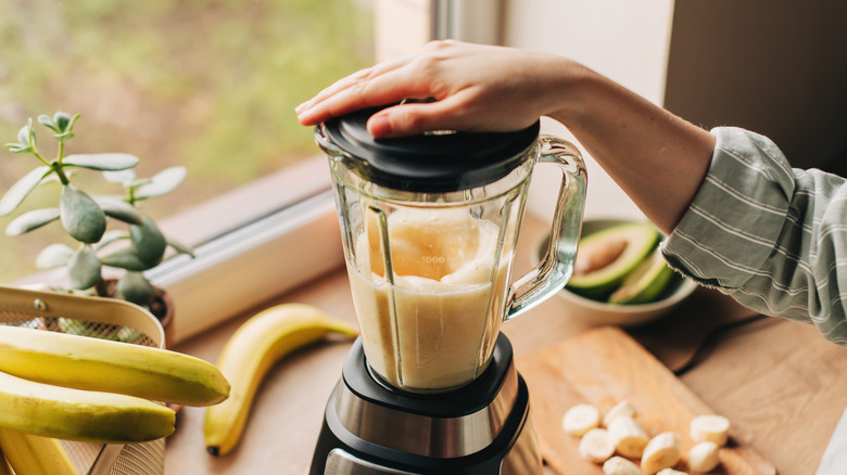 A person holding the lid on a blender while it mixes a smoothie with banana and avocado