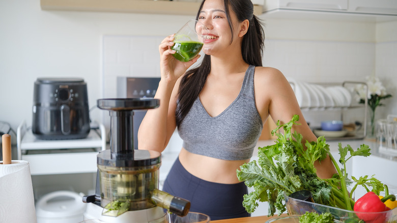 A woman drinking green juice standing next to a juicer and a container full of fruits and veggies