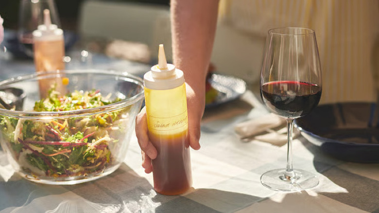 Someone holding an IKEA squeeze bottle filled with salad dressing next to a salad bowl and a glass of red wine on a dining table