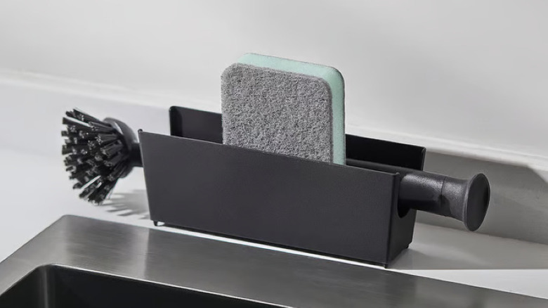 A black IKEA sink organizer with a sponge and dish brush propped on a countertop beside a sink