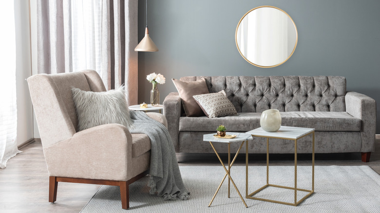 A cream armchair with grey throw and fluffy cushion with a grey sofa and cushions behind it. The wall is grey and there is a large circular mirror as well as two small tables in front of the the sofa.