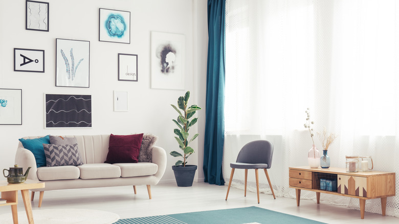 A gallery wall in a living room with huge floor to ceiling window. The pictures are hung above a cream sofa and there is also a modern chair and wooden low sideboard, along with a large rug on the floor.
