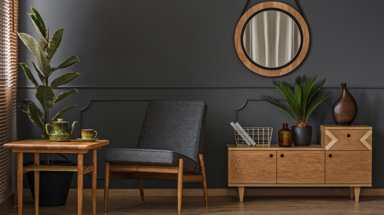 A grey walled living room with circular mirror on the wall, a g-plan style sideboard, grey armchair, coffee table and plants.