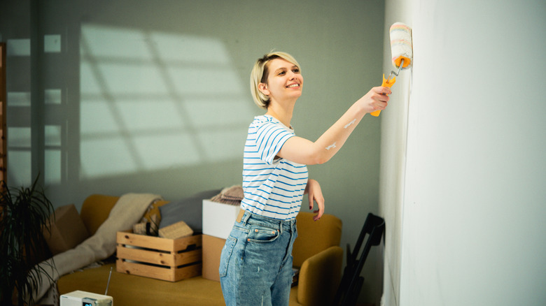 A woman using a roller to paint the wall in her living room. There is a yellow sofa with crates and boxes on it behind her.