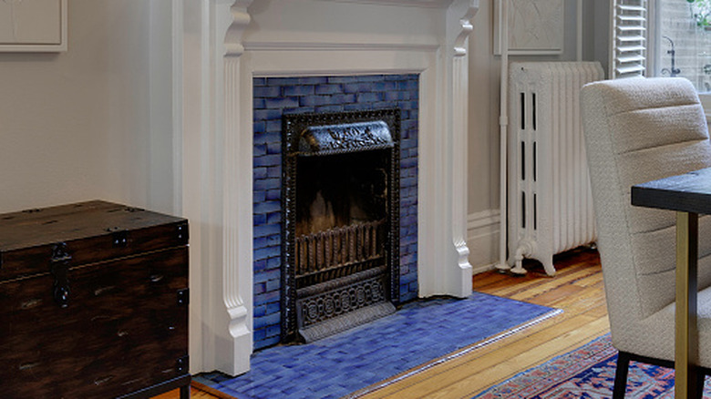 A blue tiled fireplace with an antique chest next to it. The corner of a dining table and a cream padded dining chair are also visible.