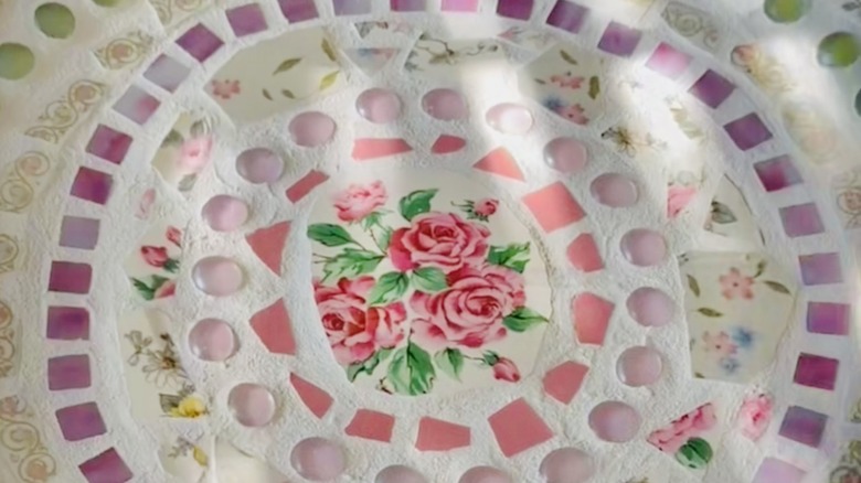 A mosaic birdbath in soft pink and lavender colors with a rose image at the center from broken china.