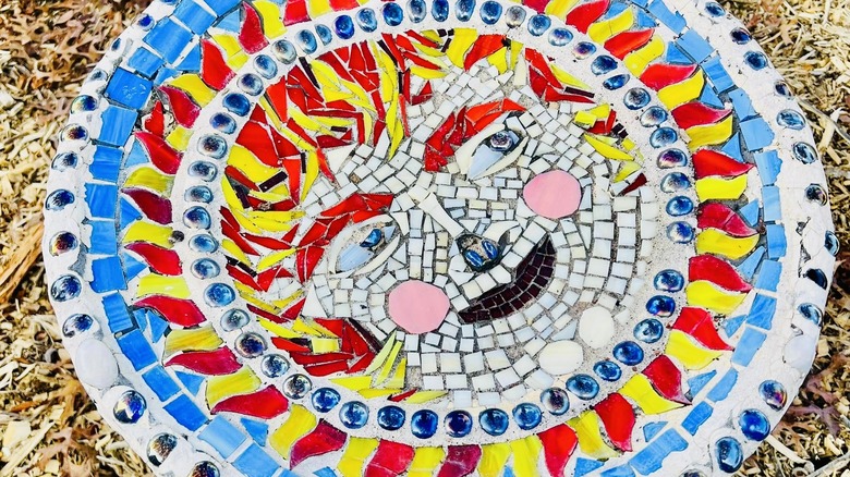 Mosaic bird bath in bright colors and broken china formed into a sun with a face.