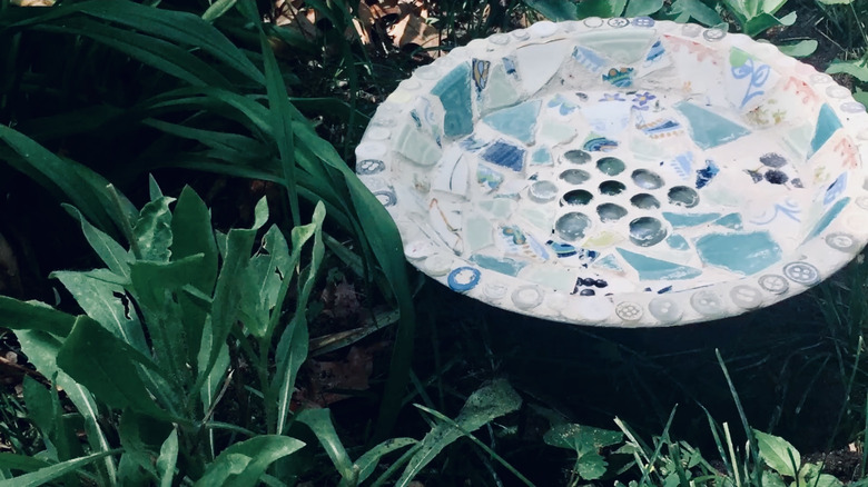 A mosaic bird bath with broken china and glass stones amid lush plantings.