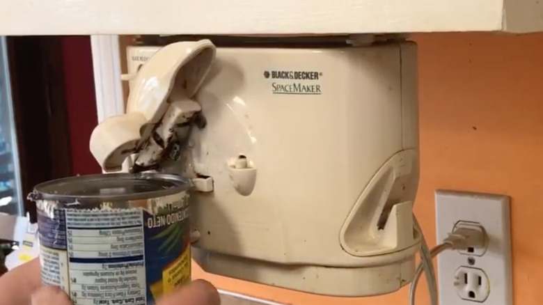 A person uses an electric can opener mounted underneath an upper kitchen cabinet.