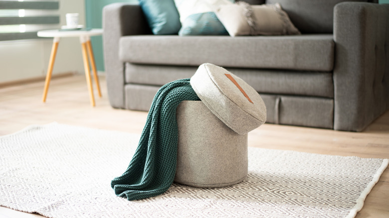 A small felt ottoman with green throw sticking out of it  and a blurred gray sofa with green and cream pillows behind it.