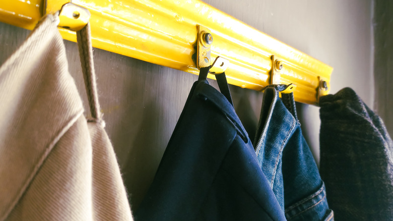 A camel coat, blue coat, denim jacket, and tweed coat hanging on brass coat hooks with a yellow board to hold them up.