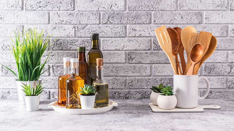 A tray with various cooking oils next to crock filled with wooden utensils against a gray brick backsplash on a kitchen counter