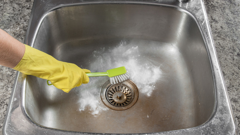 A yellow gloved-hand scrubbing a stainless steel kitchen sink with a green dish brush