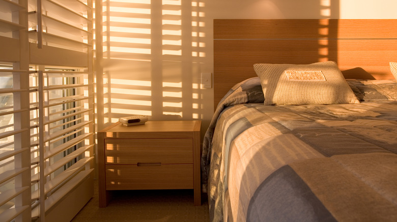 Sunlight filtering through blinds into a bedroom wiith a pine headboard and brown checked bed linen.
