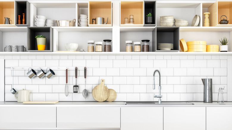 Open kitchen cabinets storing cups, plates, and canisters above a white countertop and sink