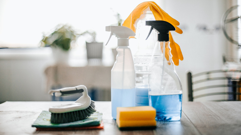 Household cleaners, a sponge, gloves, and scrub brush on a wooden table