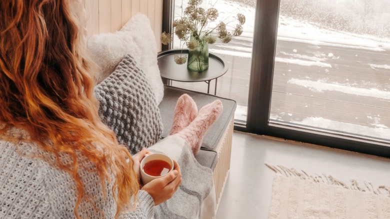 A woman sitting on a couch with tea watching it snow