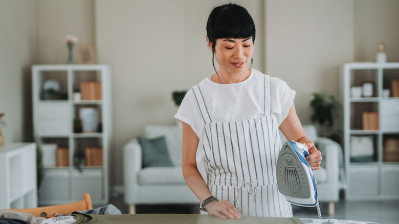 A woman at an ironing board is holding up a clean iron prior to using it