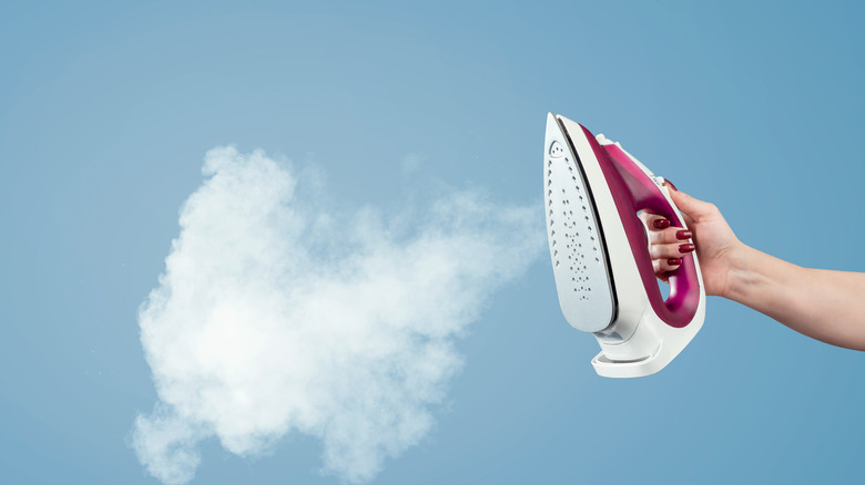 A woman's hand is holding a pink and white iron which is emitting a cloud of steam against a blue background