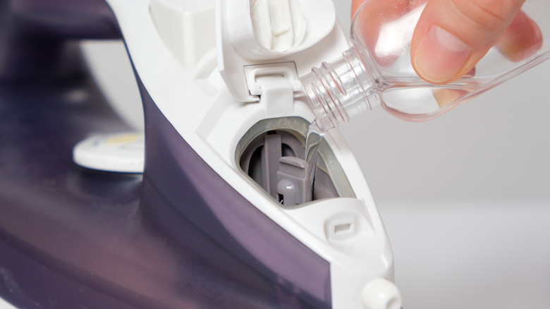 A hand is pouring a commercial product into the water reservoir of a purple and white clothes iron
