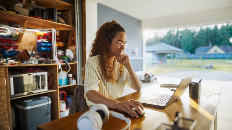 Young woman browsing laptop in cluttered garage with door open