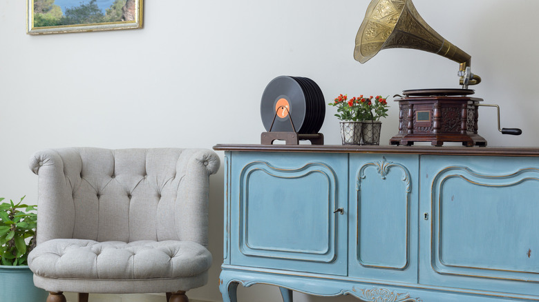 Vintage interior of retro off white armchair, vintage wooden light blue sideboard, old phonograph (gramophone) and vinyl records on background of beige wall, tiled porcelain floor, and red carpet.