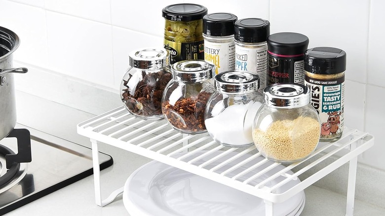 A white shelf riser on a kitchen counter with spices on top and plates underneath.