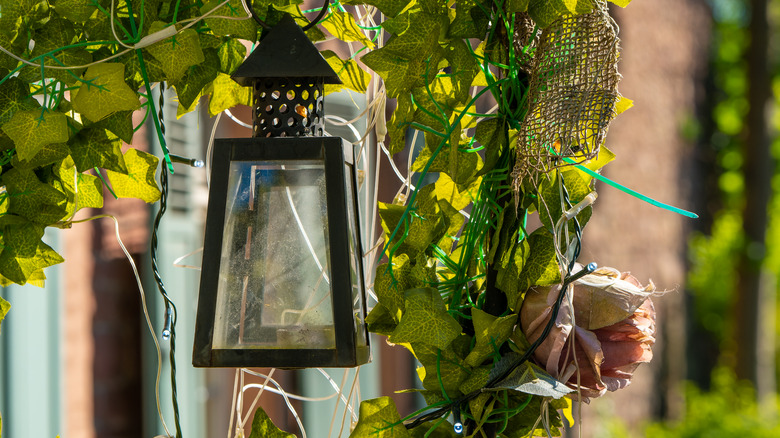 A metal carriage lantern hangs from a garden arch with faux ivy and fairy lights wrapped around it.