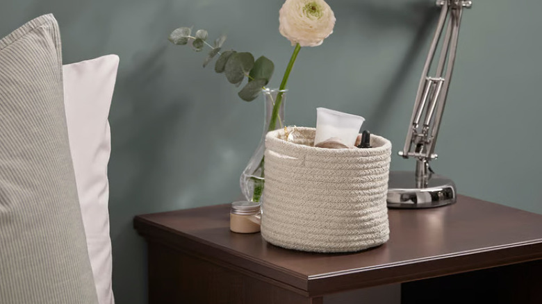 Woven organizer basket on wood nightstand with flowers, lamp, and small candle