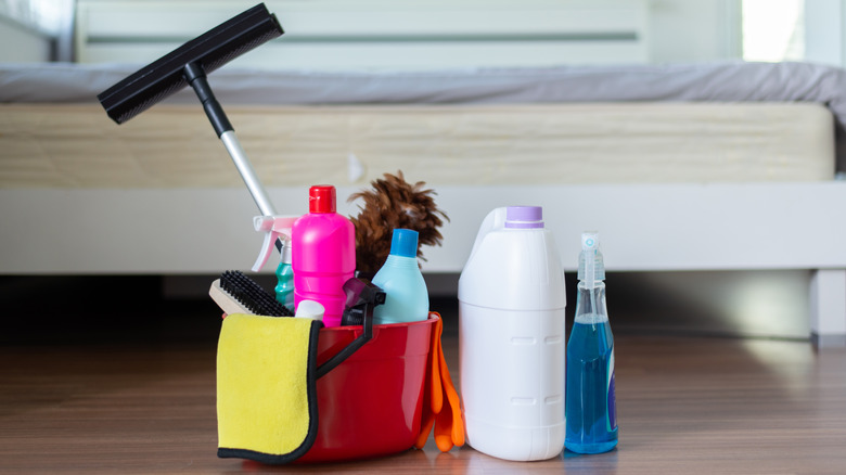 cleaning solutions, brush, squeegee, microfiber cloth, and duster in a bucket on a bedroom floor