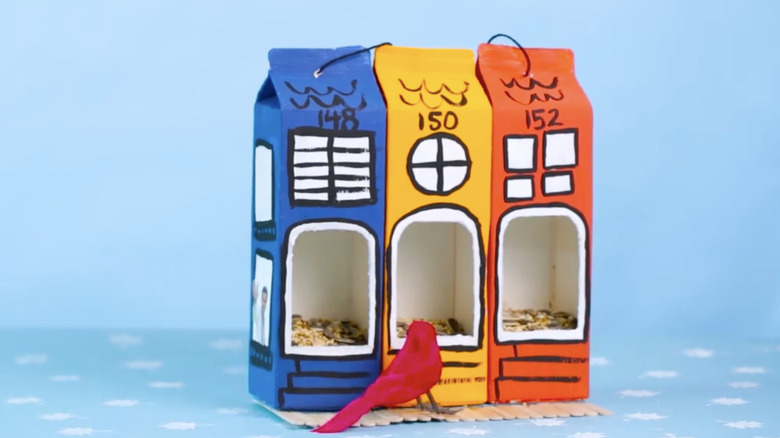 Three milk carton bird feeders are attached side by side and painted blue, yellow, and red.