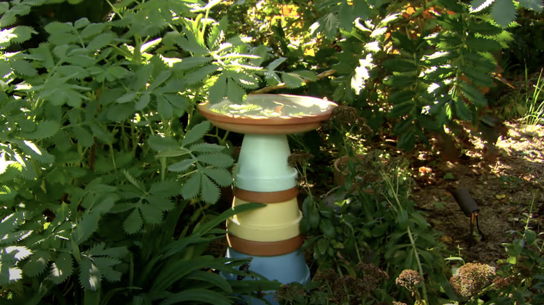 A bird bath made from painted plastic nursery pots and a saucer sits in an overgrown garden bed.