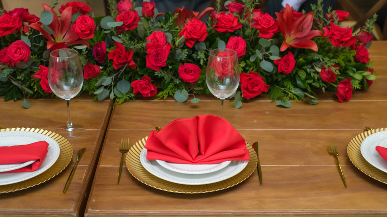 Place settings with golden chargers, white plates, and red napkins with a red rose display in the background