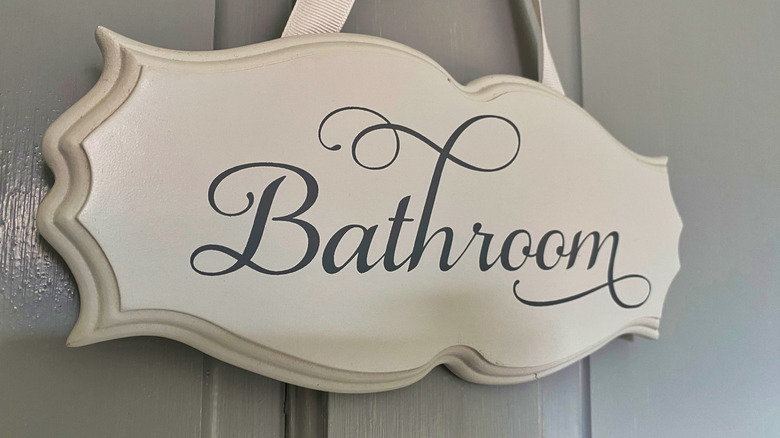 Sign reading Bathroom on wooden plaque, hanging from ribbon on gray door
