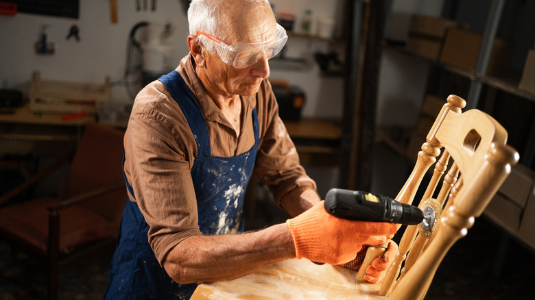 Older man sanding a wooden chair with a drill fitted with a sanding attachment