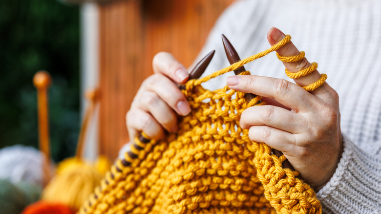 Person knitting a mustard yellow piece with two large knitting needles