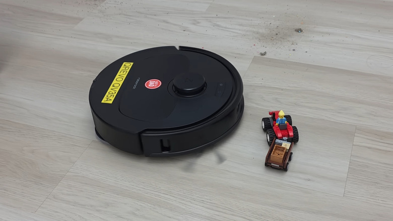 Roborock robot vacuum operating on wood floor around scattered toys and dirt