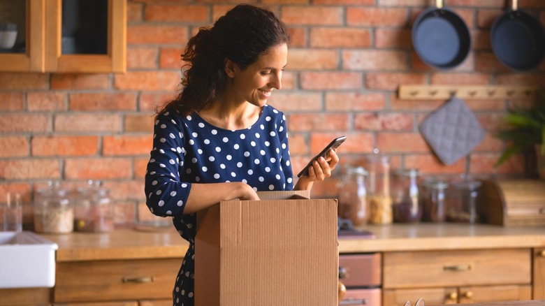 Smiling woman looking at smartphone as she unboxes new appliance in rustic kitchen